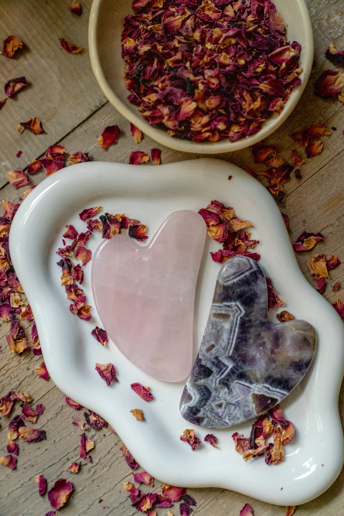 Heart-shaped pink stone and amethyst crystal on a white dish with dried rose petals.