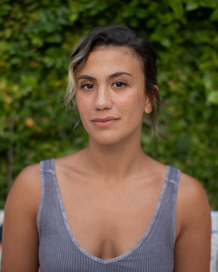 A woman with dark hair pulled back sits centered in front of a green hedge, wearing a gray ribbed tank top. Colorful outdoor pillows are visible on a white sofa behind her.
