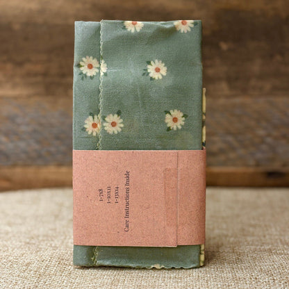 A folded fabric item, likely a pouch or small bag, wrapped in sage green cloth printed with small white daisies and secured with a dusty pink paper band labeled 'Care Instructions Inside,' set against a rustic wood and burlap background.
