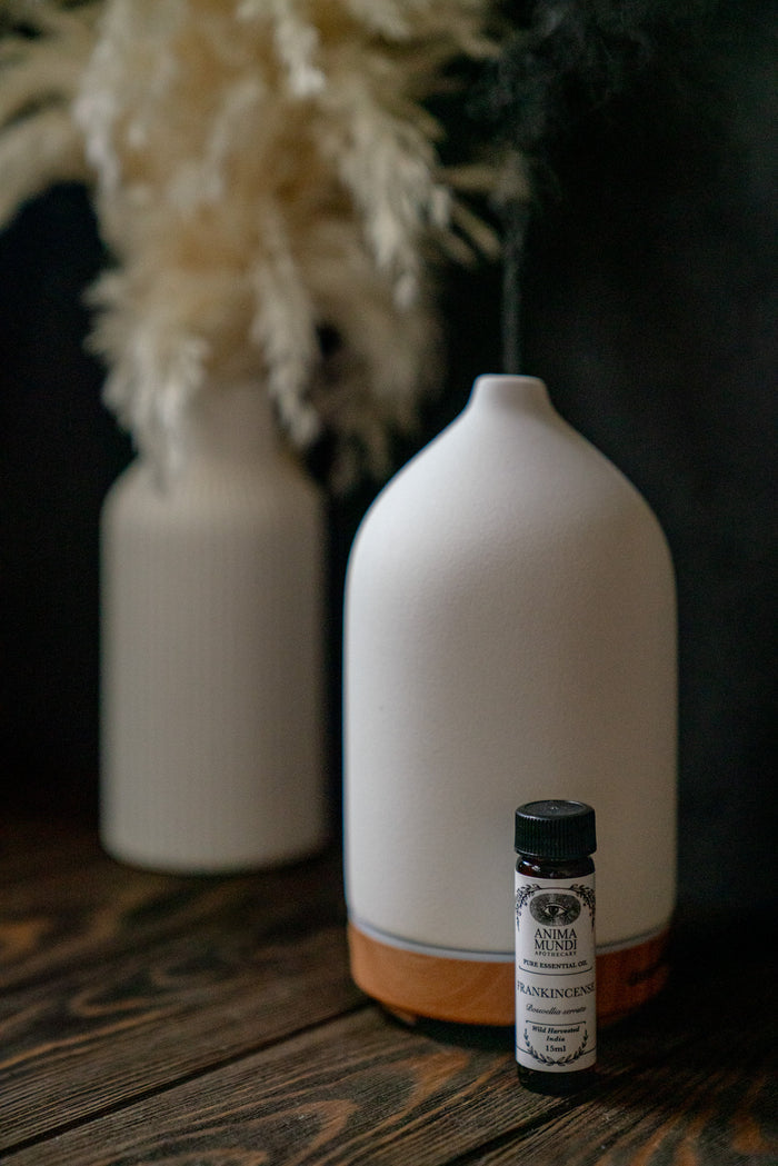 White ceramic diffuser with a small bottle on a wooden surface, with blurred white flowers in the background.
