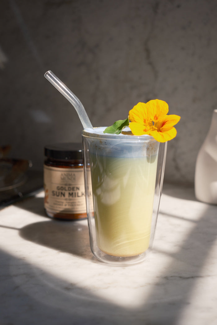 Green smoothie in a clear glass with a yellow flower, surrounded by a jar of Golden Sun Milks on a marble surface.