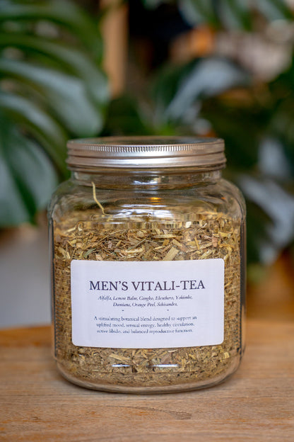 Jar of Men's Vitali-Tea with a label on a wooden surface, blurred green plants in the background