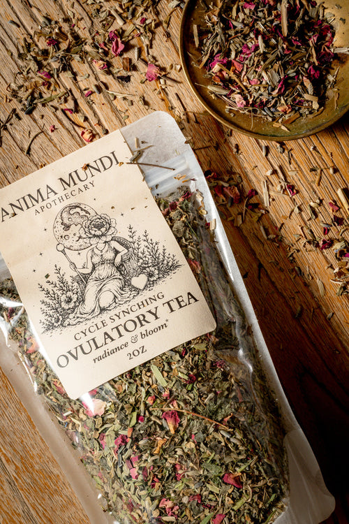 A clear plastic bag of Anima Mundi Apothecary Cycle Syncing Ovulatory Tea filled with dried herbs and pink petals, placed on a rustic wooden surface with a small dish of loose tea nearby.