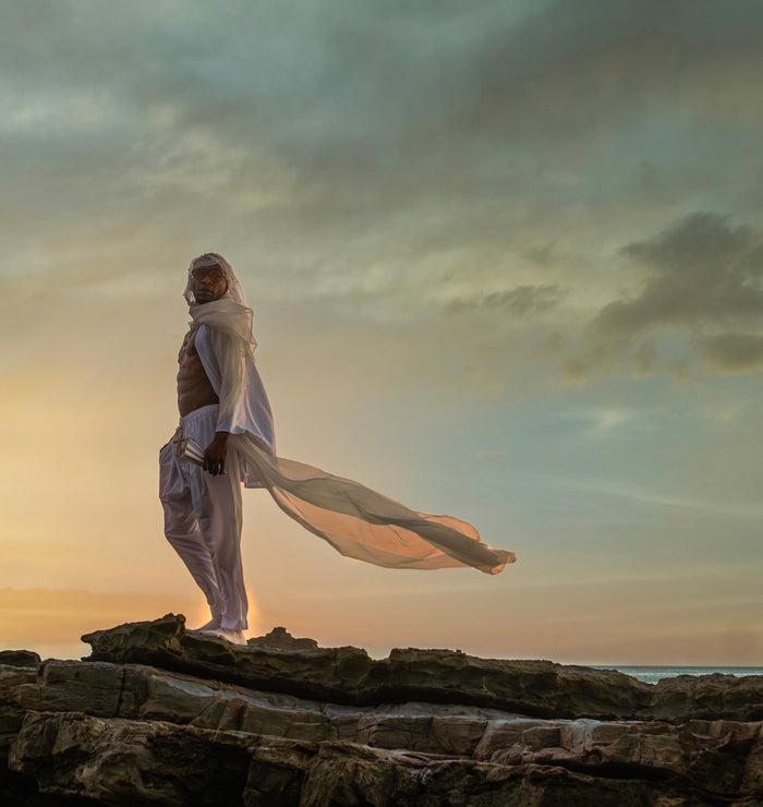 Person standing on a rocky outcrop with a flowing garment against a dramatic sky.