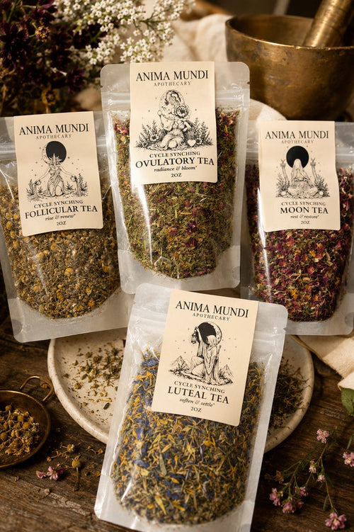 Three clear stand-up pouches of Anima Mundi botanical tea arranged on a rustic wooden surface with floral props and brass kitchenware. The visible labels read Extra Virgin Ovulatory Tea, Follicular Tea, and Luteal Tea.