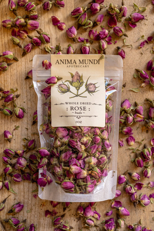 Packaged dried rose buds with 'Anima Mundi Apothecary' label on a wooden surface with rose buds scattered around.